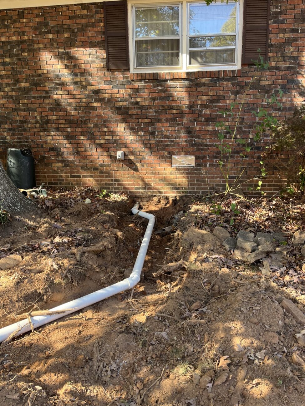 A white PVC pipe is partially buried in a trench running from a brick house wall, with scattered soil and leaves around. A window with shutters is visible on the building above.